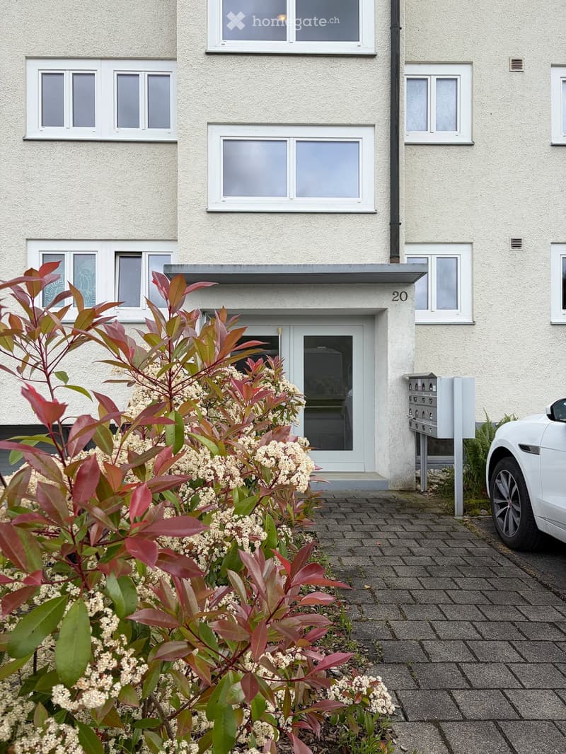 Two-story residential building with white facade, multiple windows, and a small garden area with flowering shrubs in front of the entrance.