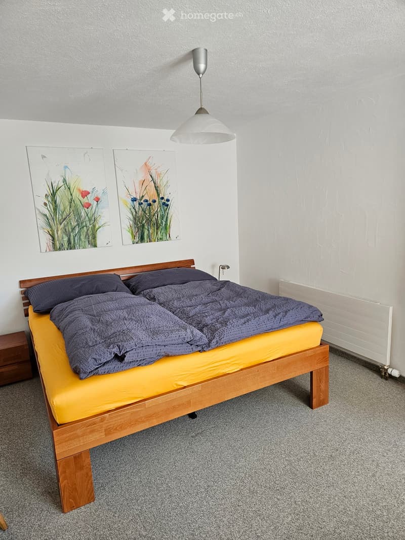 A neatly arranged bedroom with a wooden bed, yellow mattress, dark blue duvet, and matching pillows. A pendant light hangs above the bed, and two abstract paintings adorn the wall.
