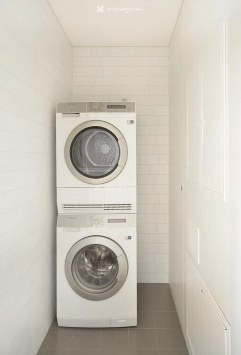 A compact, modern laundry room with a stacked washer and dryer unit, white tiled walls, and a dark tiled floor.
