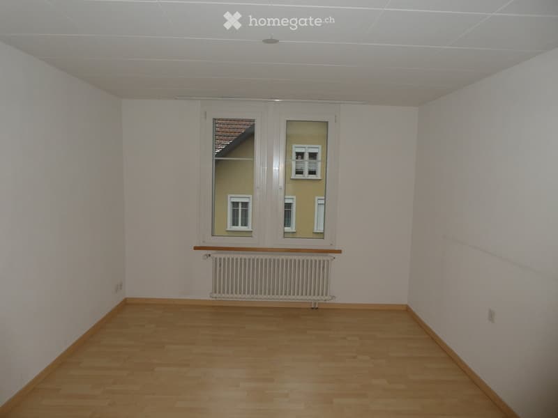 Empty room with light wooden flooring, white walls, and a window with a radiator below it.