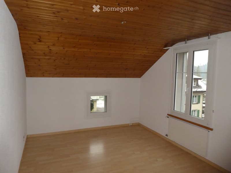 Empty room with wooden flooring, white walls, and wooden ceiling. Two windows provide natural light, and a radiator is visible on one wall.