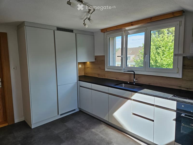 Modern kitchen with white cabinets, dark countertops, and a stainless steel sink and oven. Large windows provide natural light.