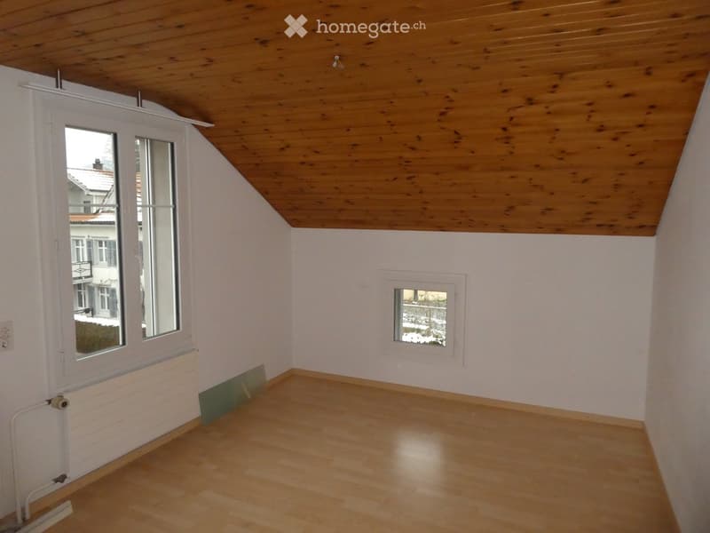 Empty room with wooden floor, white walls, wooden slanted ceiling, large window, and small window.