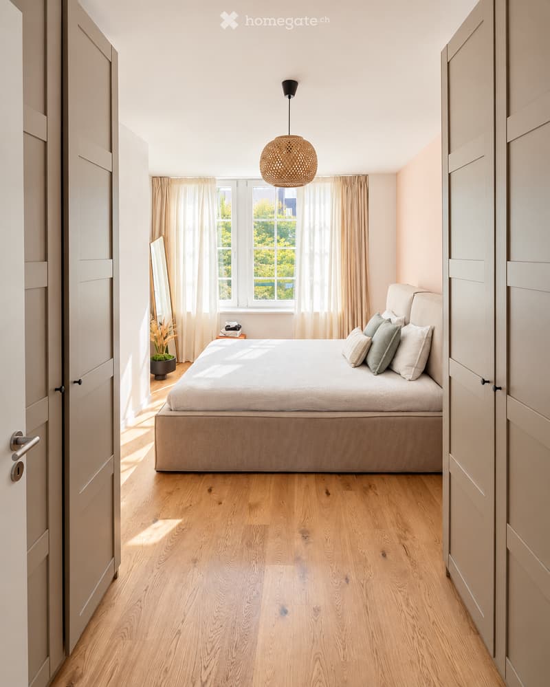 Modern bedroom with light wooden flooring, a large bed with pillows, a nightstand with a mirror, and a hanging woven light fixture.