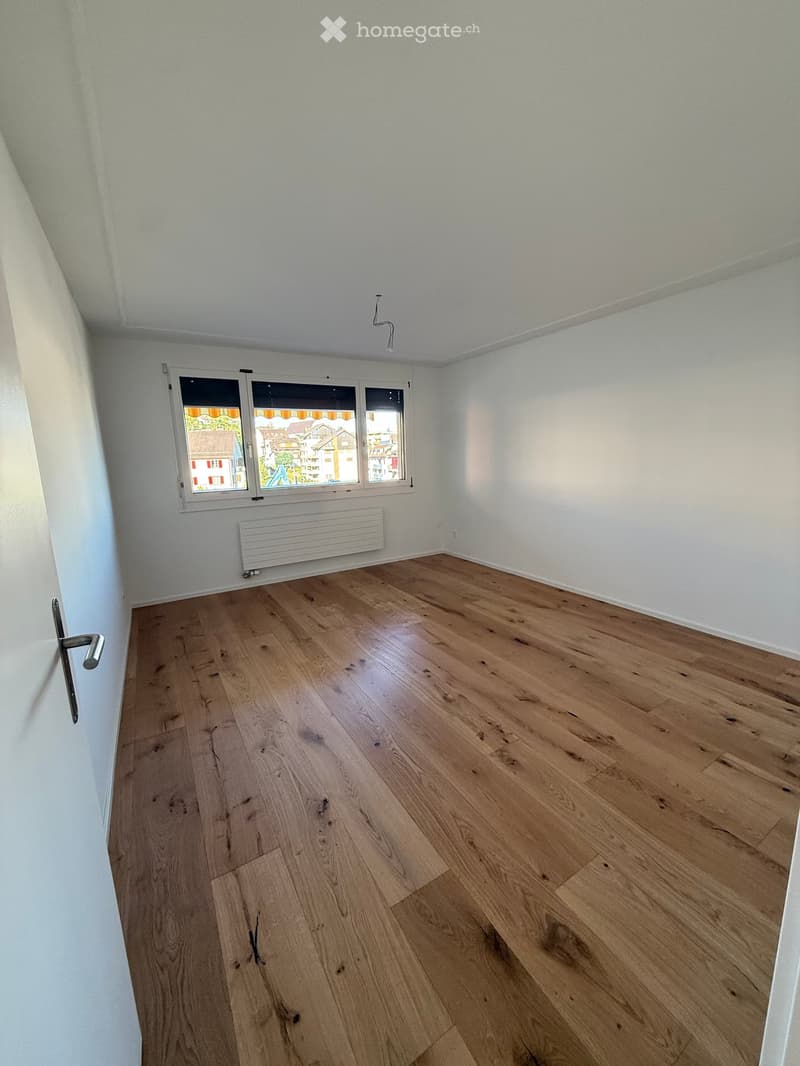 Empty room with light wooden flooring, white walls, and a large window with a view of the outside. A radiator is visible below the window, and a door is on the left side.
