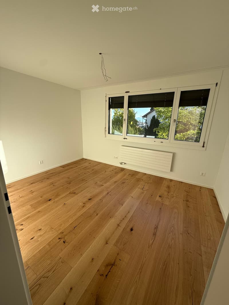 Empty room with light wooden flooring, white walls, and large windows with blinds. A white radiator is mounted on the wall below the windows.