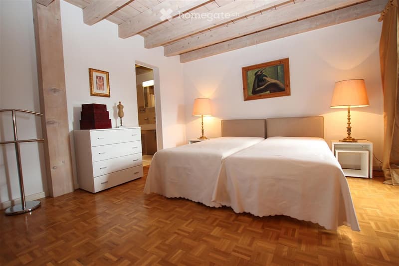 Cozy bedroom with wooden herringbone flooring, a large bed with white bedding, two nightstands with lamps, a dresser with drawers, and a mirror in the adjacent bathroom.