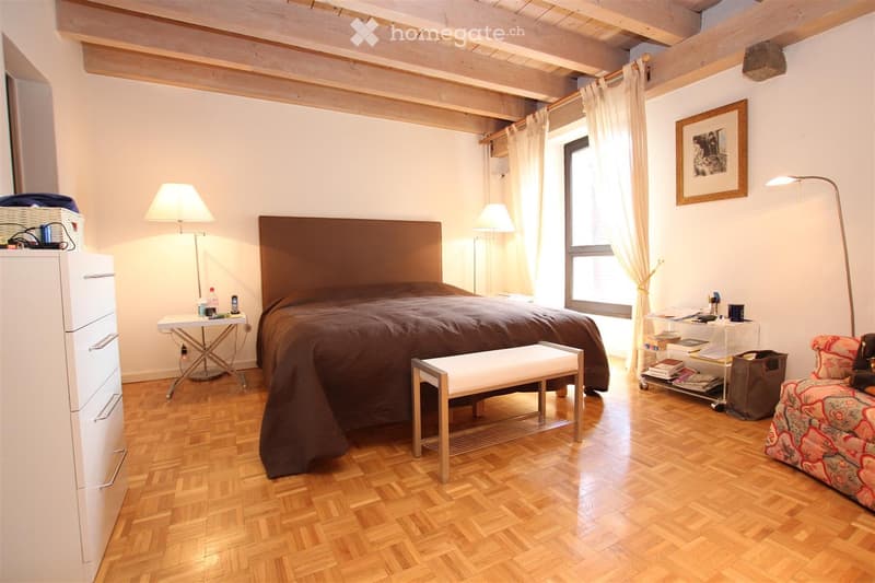 Cozy bedroom with wooden flooring, a large bed with a dark brown headboard and matching duvet, a white bench at the foot of the bed, a patterned sofa, a nightstand with a lamp, a small table with a lamp, and a drawer unit.