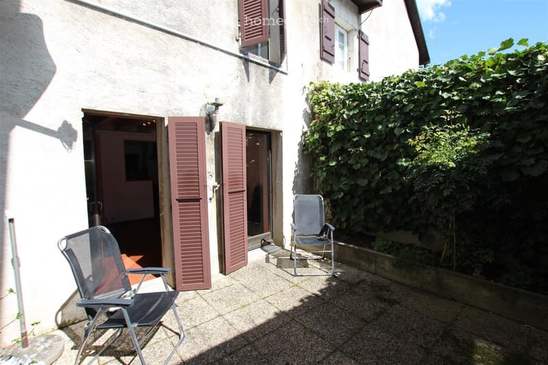 A small outdoor patio area with two folding chairs, a building entrance with shutters, and a lush green hedge.