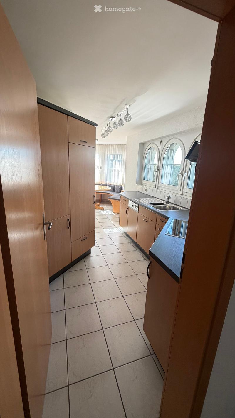 A modern kitchen with wooden cabinets, tiled floor, and a sink area. A small living area with a sofa and table is visible through an open doorway.