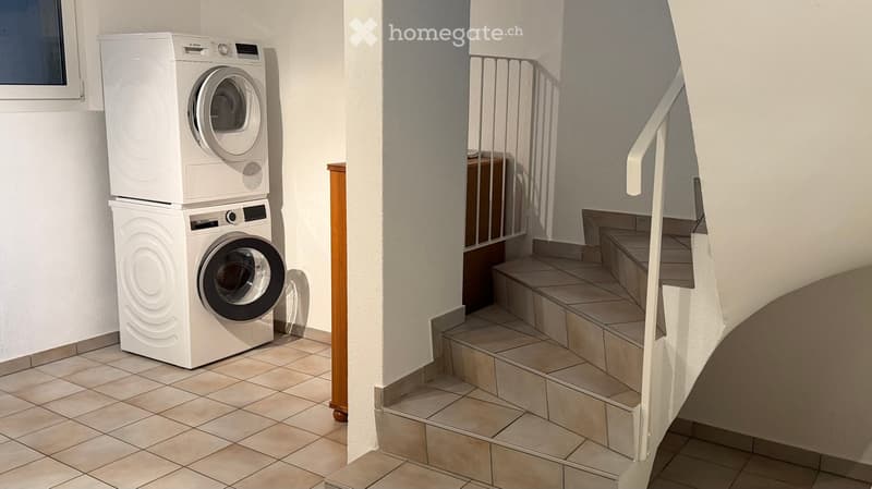 Hallway with tiled floor, white walls, staircase with metal railing, and stacked washing machines in the corner.