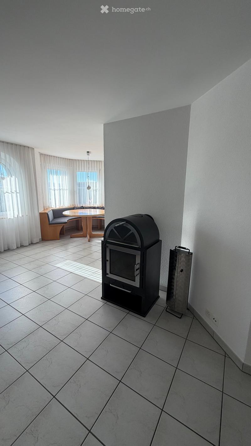 A modern room with a tiled floor, a fireplace, a round wooden table with a bench, and large windows with white curtains.
