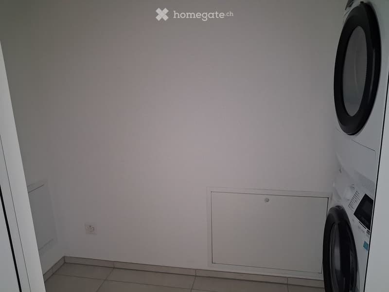 A small bathroom with a washing machine placed against the wall, light-colored tiled floor, and white walls.