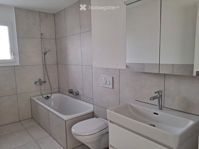 Modern bathroom with a bathtub, toilet, sink, and showerhead. Light gray tiles and white fixtures.