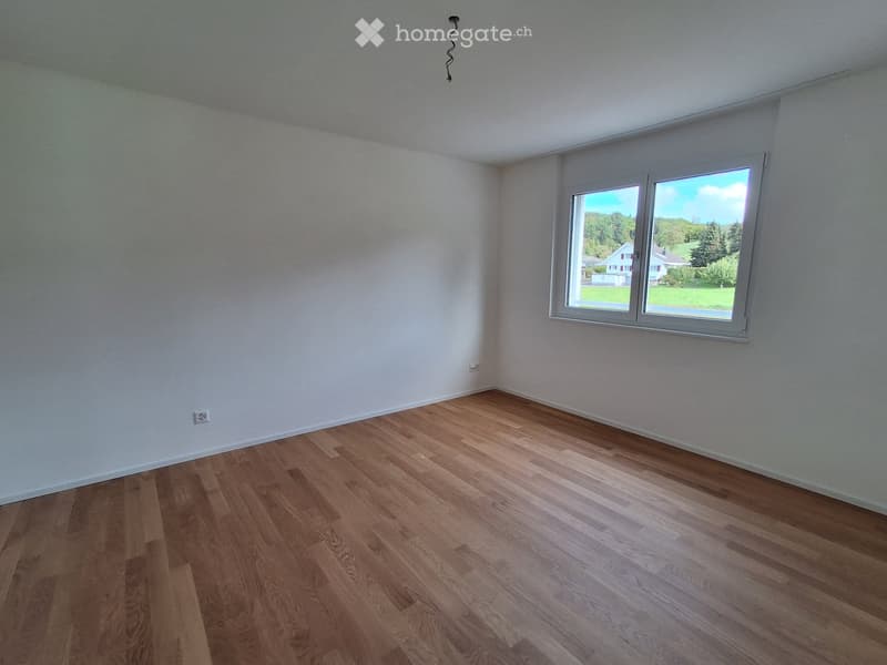 Empty room with wooden flooring and white walls, featuring a large window with a view of greenery and houses outside.