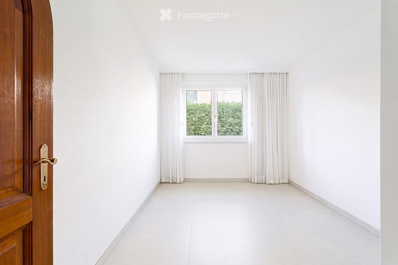Empty hallway with white tiled floor, white walls, and a wooden door on the left. A window with white curtains is visible at the far end, showing greenery outside.