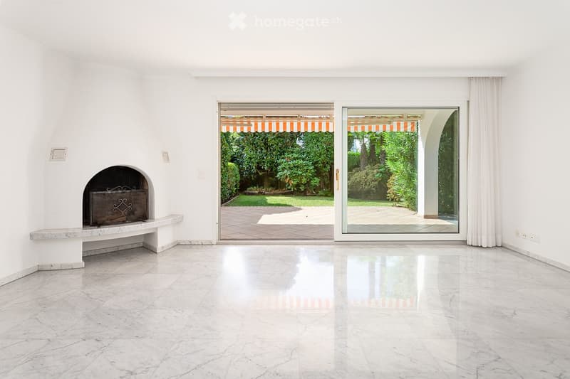 Empty room with white walls, marble floor, fireplace, large sliding glass doors leading to a garden.