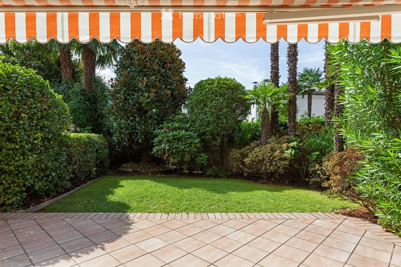 A well-maintained garden with a tiled patio, lush greenery, and a striped awning providing shade.