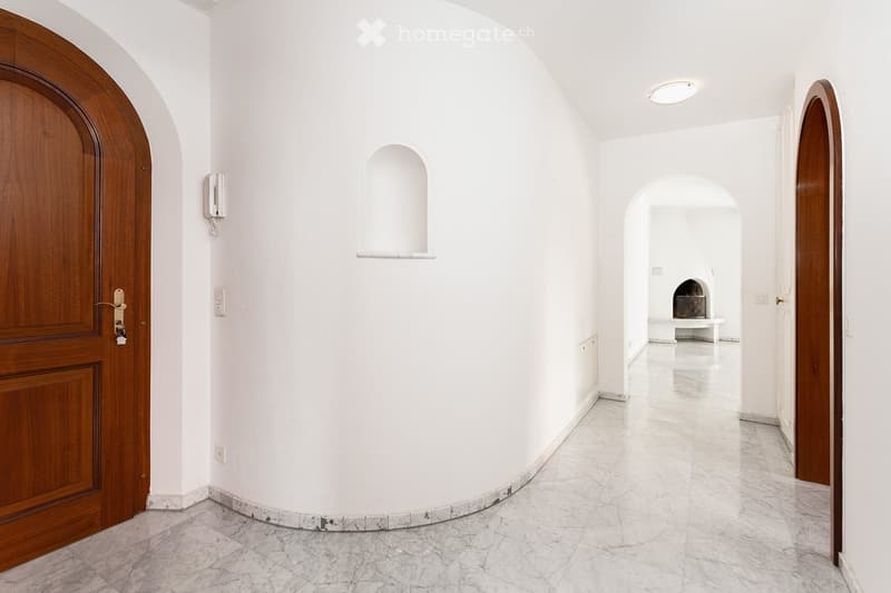 Empty hallway with white walls, marble flooring, wooden doors, and recessed lighting. Features arched doorways and a small niche with a shelf.