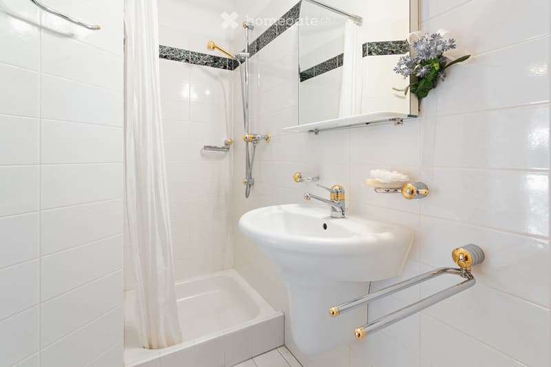 Modern bathroom with white tiles, a sink, bathtub, shower curtain, and a decorative flower arrangement on the wall.