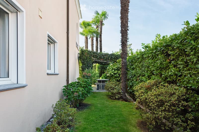A well-maintained garden area with neatly trimmed grass, various shrubs, and palm trees, featuring a small seating area with a table and chairs.