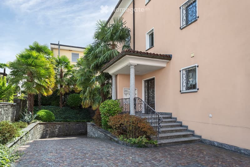 Two-story house with beige exterior, white-framed windows, decorative wrought-iron railings, and a covered entrance with columns. The house is surrounded by palm trees and shrubs, with a paved brick driveway leading to the entrance steps.