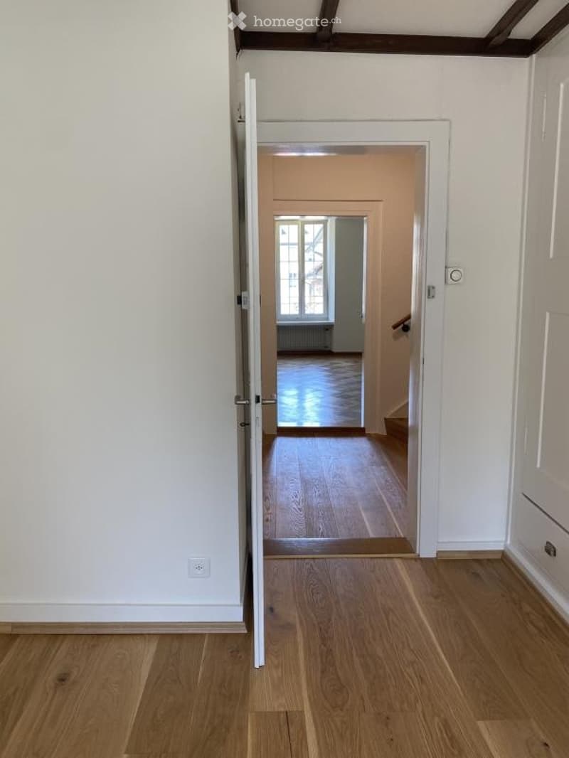 Open doorway leading to a hallway with wooden flooring and white walls, visible in the background is another room with large windows and a radiator.