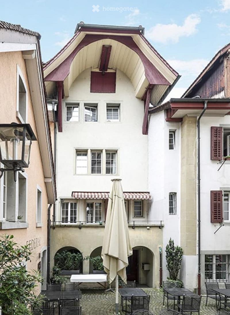 A charming multi-story building with a distinctive red and white facade, featuring arched windows and a balcony area. The ground floor includes outdoor seating with tables and chairs, and a large umbrella provides shade.