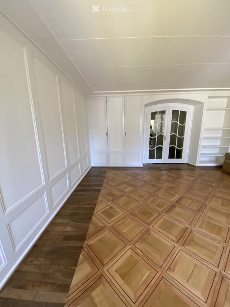 Empty room with white paneled walls, wooden floor, and a large glass door leading outside.