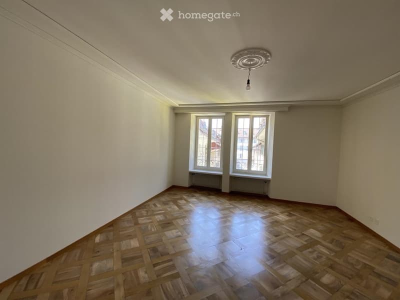 Empty room with wooden parquet flooring, white walls, and two windows with metal railings. The room has a simple ceiling light fixture and radiator under the windows.