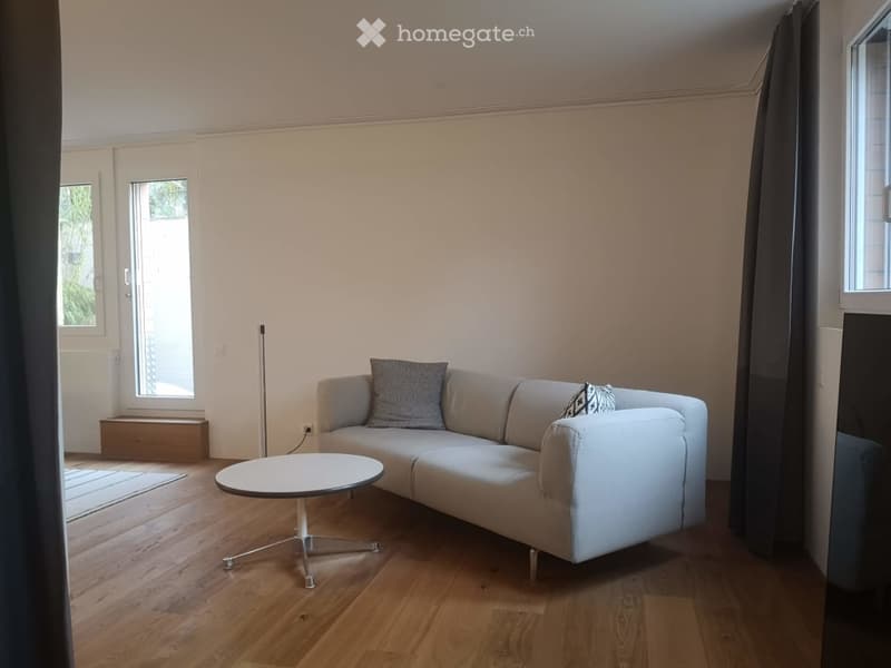 Modern living room with light-colored sofa, coffee table, and decorative cushions. The room features wooden flooring and large windows with curtains.
