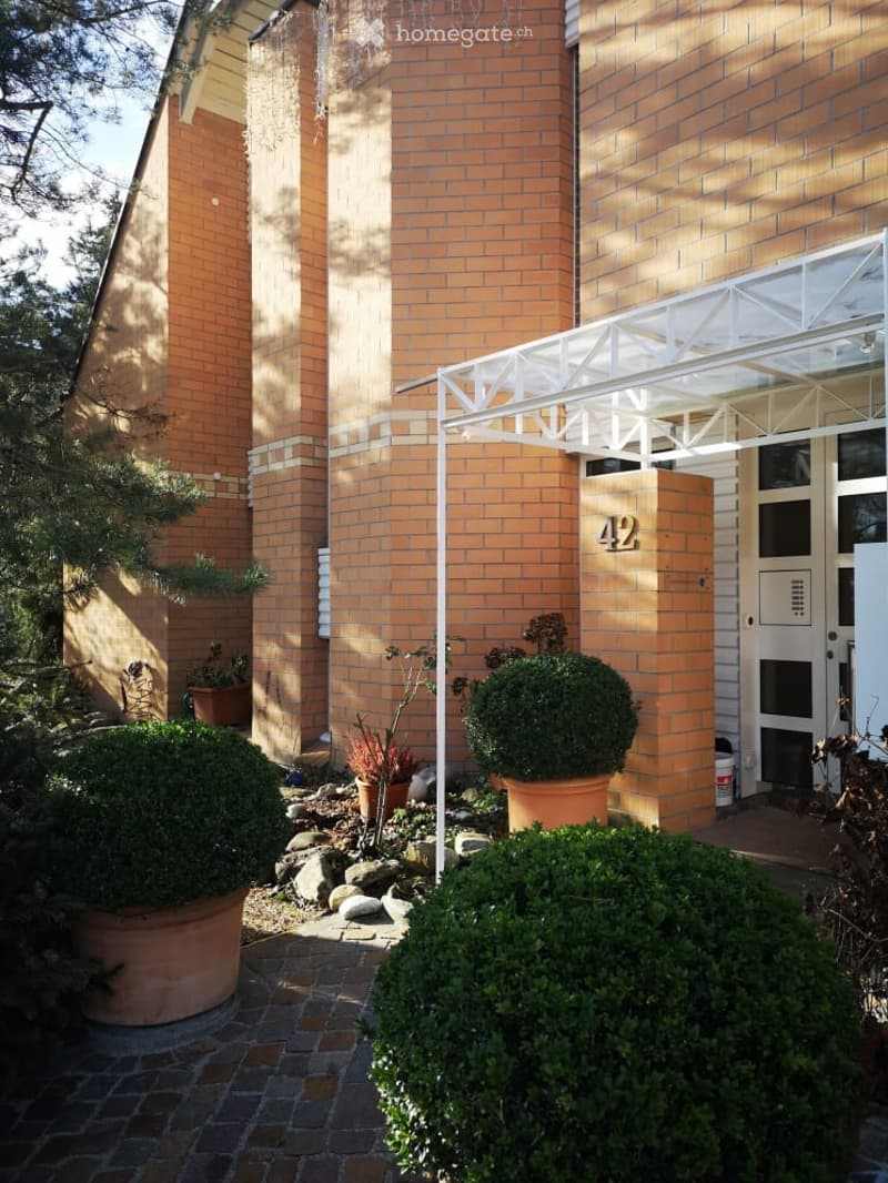 A two-story brick building with a modern entrance featuring a white canopy and the number "42" displayed prominently. The entrance is flanked by potted plants and shrubs, with a paved pathway leading to the door.
