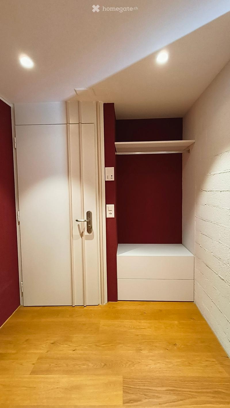 A narrow hallway with wooden flooring, white walls, and a closed white door on the left. A built-in white shelf unit is mounted on the right wall, with a maroon accent wall behind it.