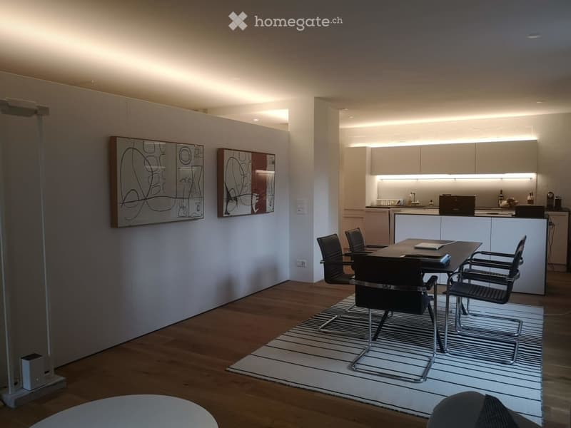 Modern, minimalist room with a wooden floor, white walls, and integrated kitchen area. Features a sleek black dining table with matching chairs, a white rug, and contemporary art on the walls. The kitchen includes white cabinets, a sink, and a refrigerato
