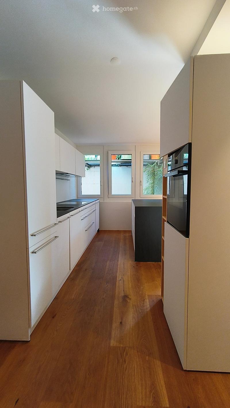 Modern kitchen with white cabinets, wooden flooring, and stainless steel appliances including a stove and oven. Three windows provide natural light.
