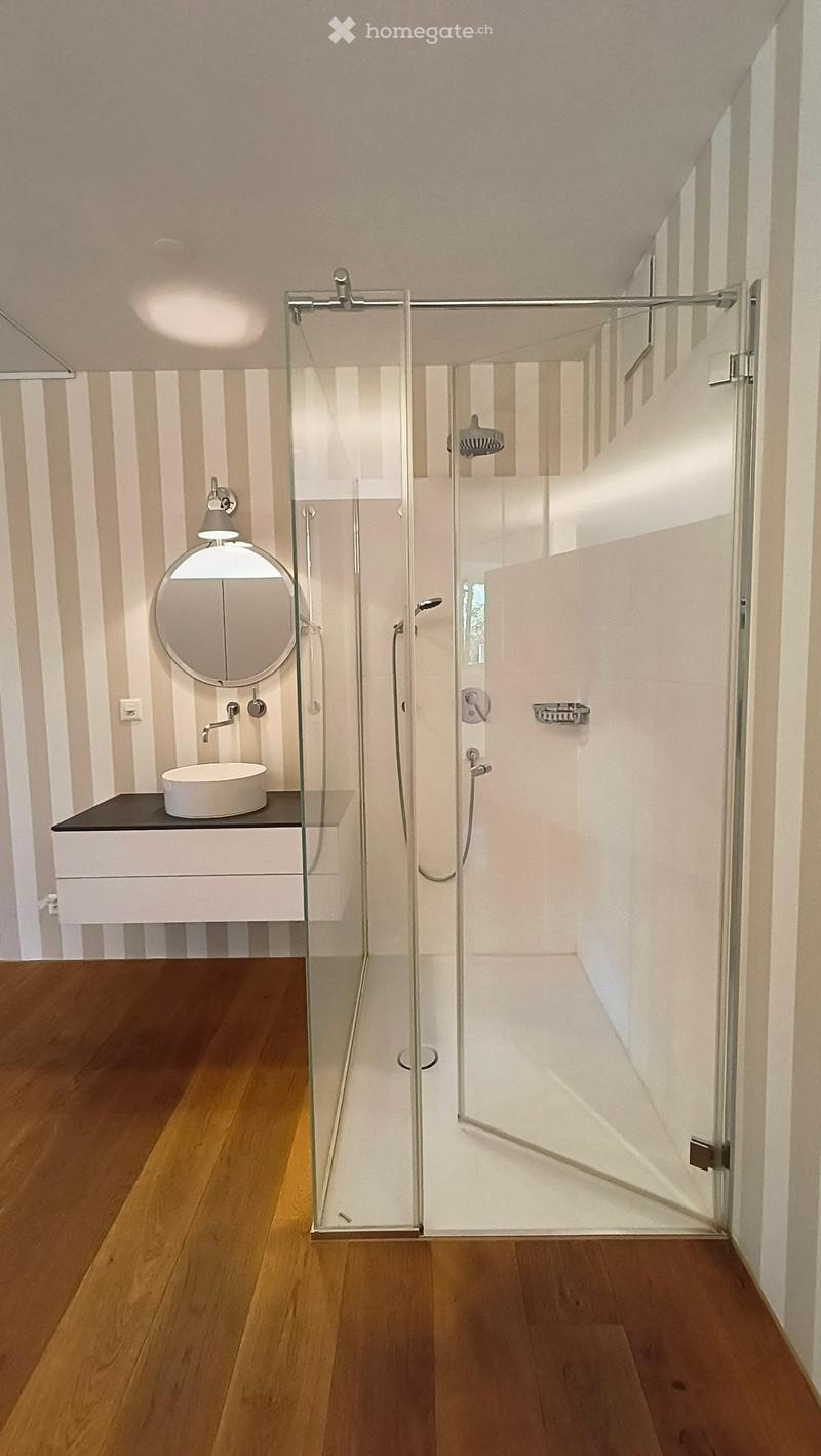 Modern bathroom with wooden flooring, a glass-enclosed shower, a white sink, and a round mirror.