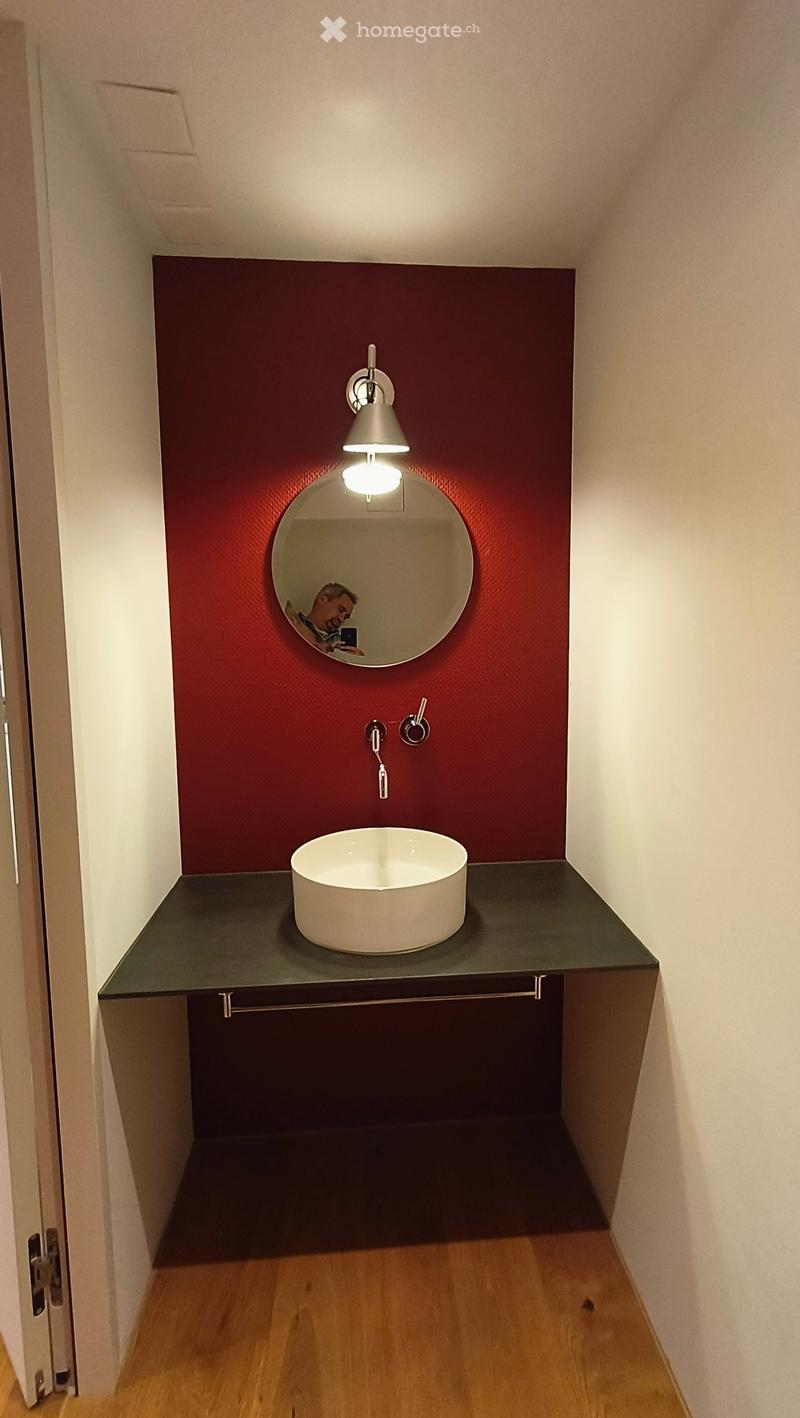 Modern bathroom with a dark green countertop, white sink, round mirror, and wall-mounted light fixture.