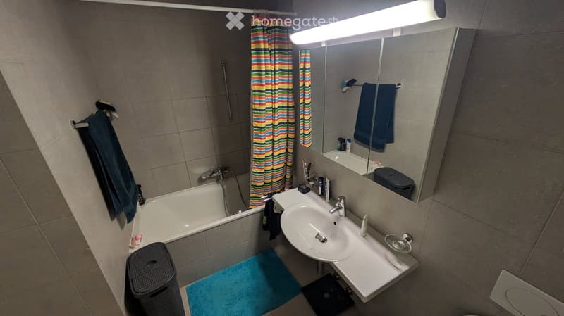 Modern bathroom with a bathtub, sink, mirror, and various accessories. Features tiled walls and floor, a colorful shower curtain, and neatly arranged towels.