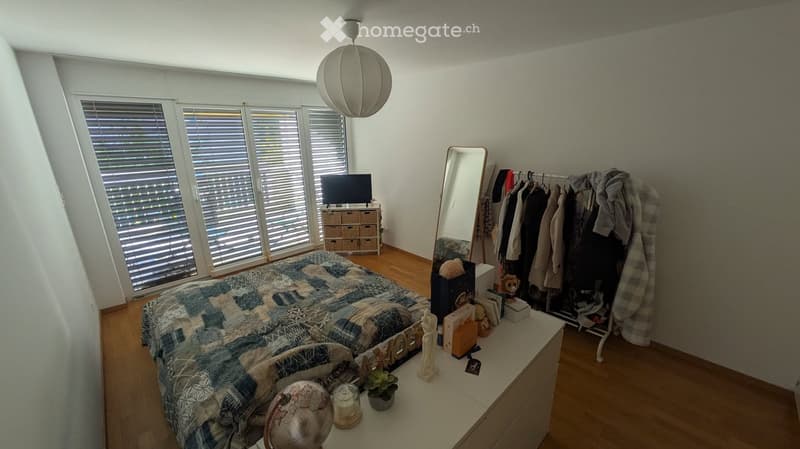 Well-lit bedroom with wooden flooring, large windows with blinds, a bed with a patterned duvet, a nightstand with decorative items, a clothing rack, a storage unit with a TV, and a mirror.