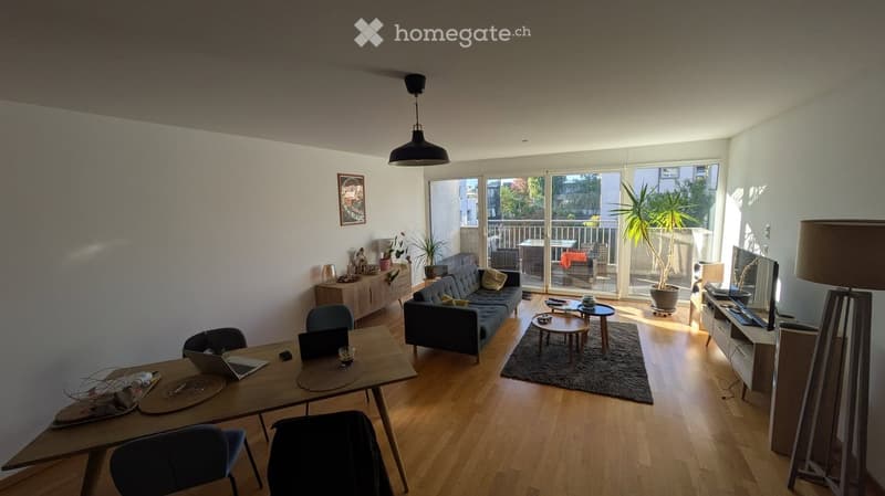 Modern living room with wooden flooring, large windows, and a balcony. Features a sofa, coffee table, dining table with chairs, laptop, and a TV stand with a screen.