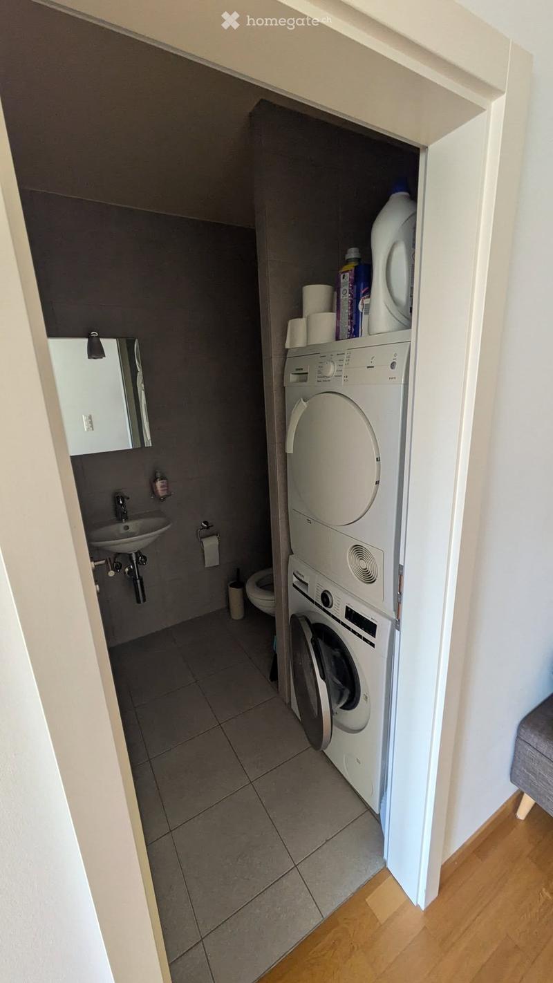 Compact bathroom with a sink, mirror, toilet, and a stacked washing machine and dryer unit. The floor is tiled, and the walls are painted in neutral tones.