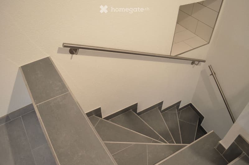 Modern staircase with grey tiled steps and a sleek metal handrail, set against white walls and a light-colored ceiling.