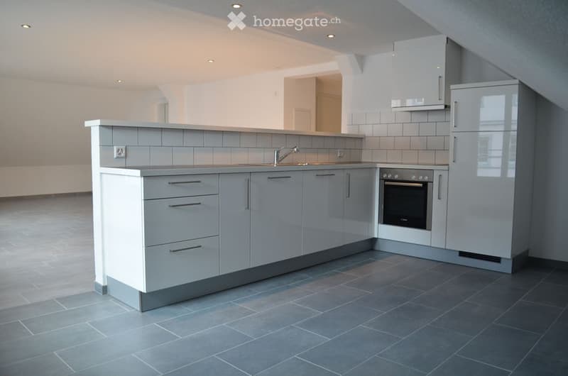 Modern kitchen with grey cabinets, a central island, a sink with a faucet, an oven, and a refrigerator. The floor is tiled in dark grey, and the walls are white with grey tiles around the sink area.