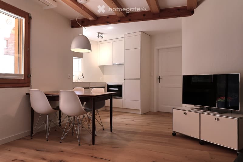 Open-plan living and dining area with wooden flooring, white chairs around a dark wooden table, a television on a cabinet, and a modern kitchen with white cabinets and an oven.