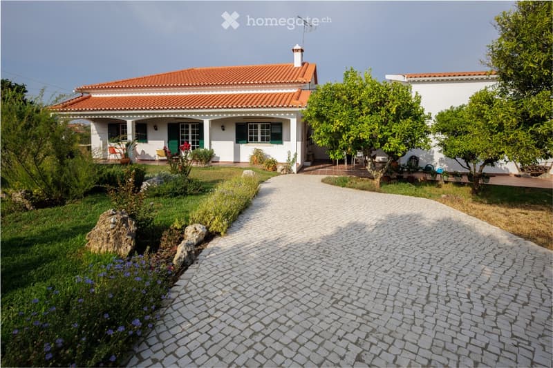 A two-story white house with a red-tiled roof, surrounded by a well-maintained garden and a cobblestone pathway leading to the entrance.