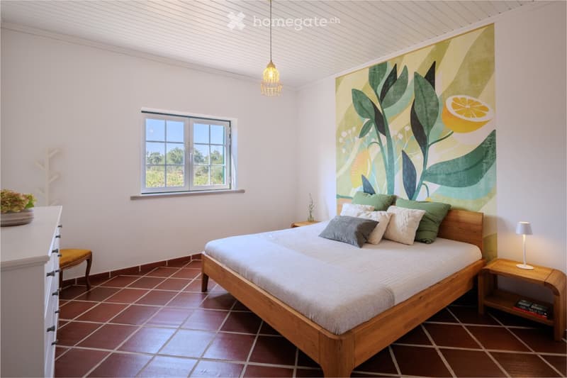 A cozy bedroom with a wooden bed, multiple pillows, a nightstand with a lamp, a small stool, a dresser with a vase, and a large window with a view.