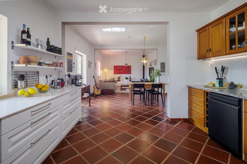 Open-plan kitchen and living area with white cabinets, granite countertops, and a tiled floor. The kitchen features a coffee maker, bottles, and a bowl of lemons. The living area includes a sofa, a coffee table, a guitar, and a dining table with chairs.
