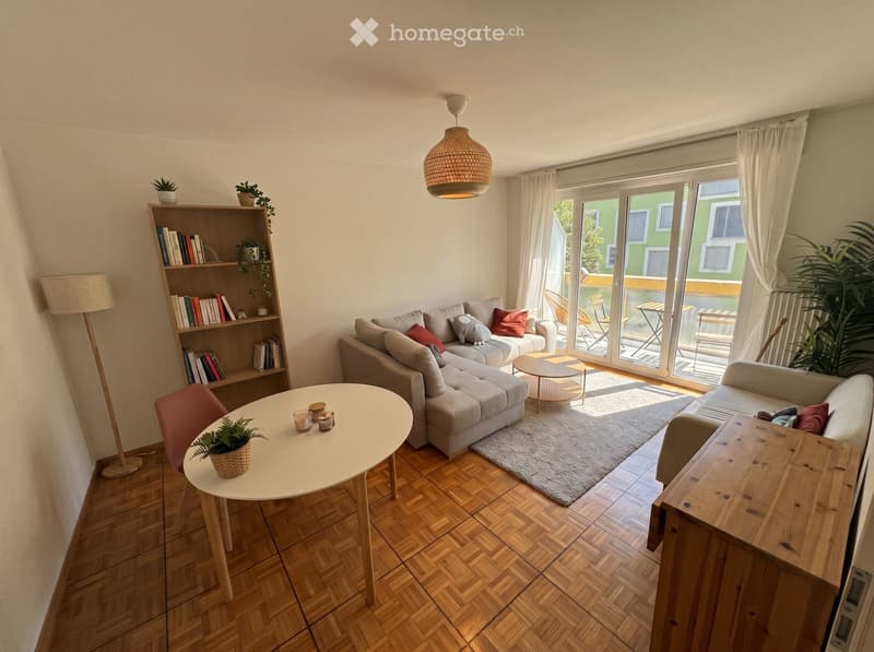 Modern living room with wooden flooring, large window, grey sectional sofa, coffee table, bookshelf, and small round table with a potted plant.