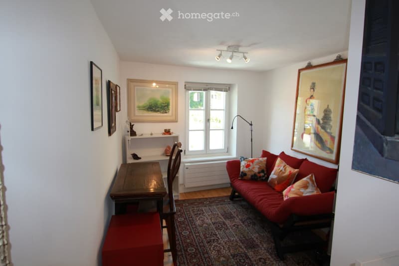 Cozy living room with a red sofa, wooden table, chair, and a colorful rug. The room features several picture frames on the walls and a window letting in natural light.