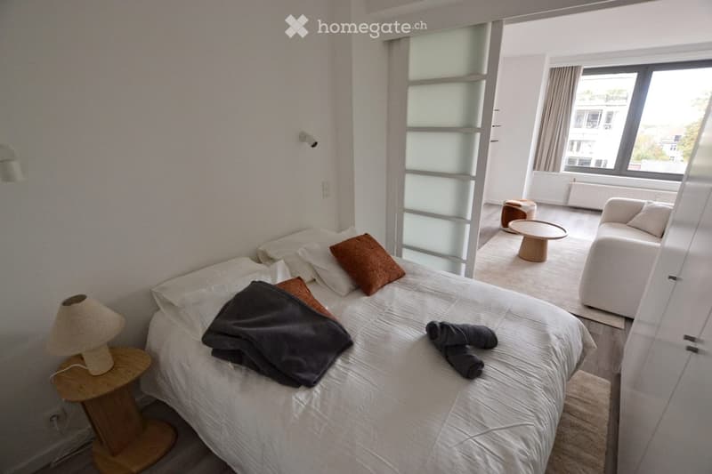 Modern bedroom with a large bed, nightstand with lamp, sofa, coffee table, and a view of the city through large windows.